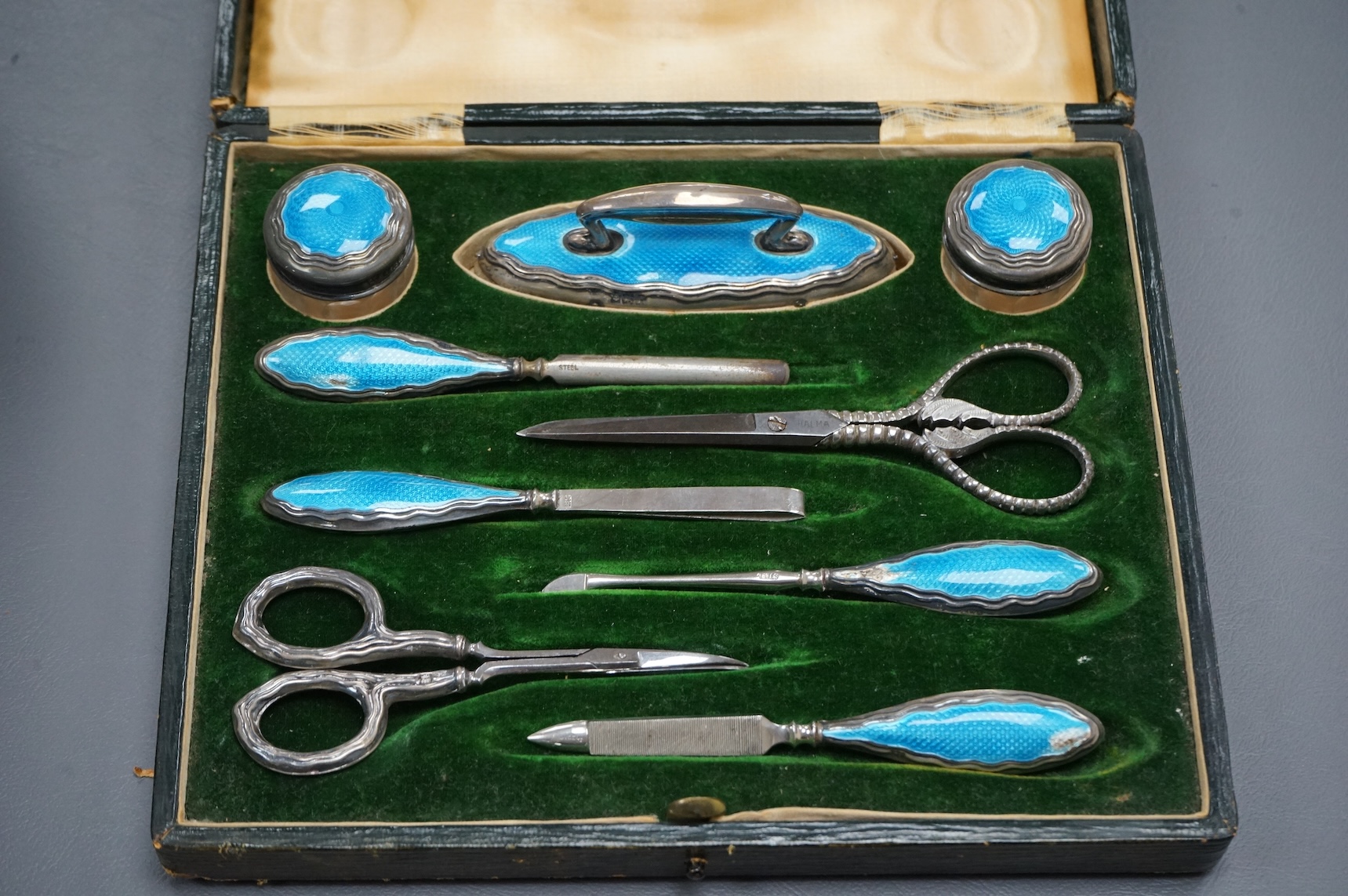 A cased George V eight piece (ex 9 piece) manicure and toilet jar set, comprising seven silver and enamel mounted items, Boots Pure Drug Company, Chester and Birmingham, 1910, together with a matching pair of steel sciss